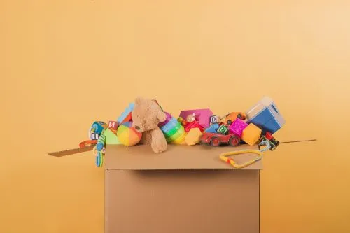 Cardboard box overflowing with colorful toys against a yellow background.