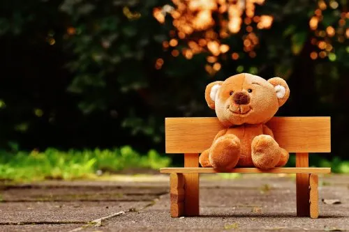 A teddy bear sits on a small wooden bench outdoors with a blurred background.