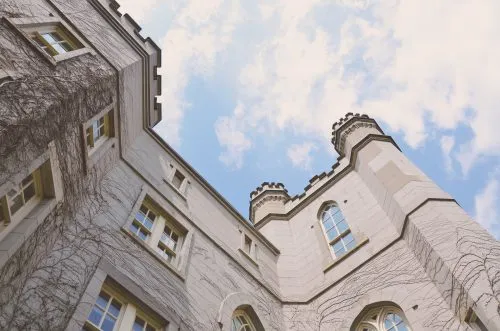 Upward view of a gray, vine-covered castle-like building against a blue sky with clouds.