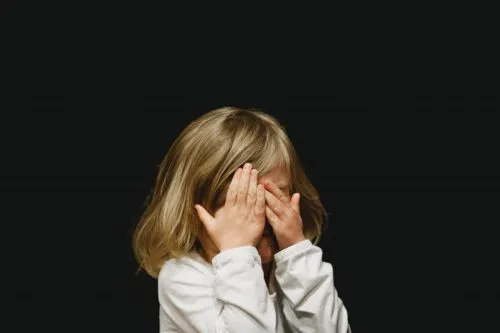 Child covering face with hands against a dark background.