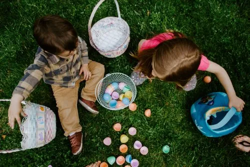 Two children sit on grass with baskets collecting colorful plastic eggs.