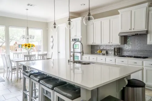 Bright kitchen with a large white island, gray backsplash, and dining area with sunflowers in a vase.