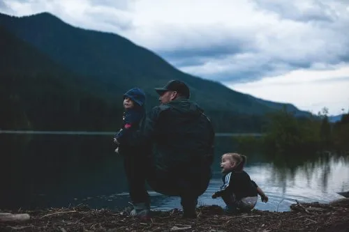 An adult and two children dressed in dark clothing kneel by a lake with mountains in the background.