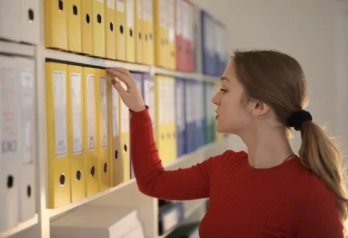 A woman in a red sweater selects a file from shelves filled with yellow binders.