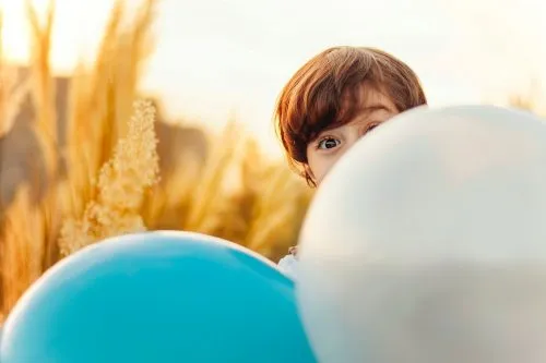 Child peeking over blue and white balloons in a sunny field.
