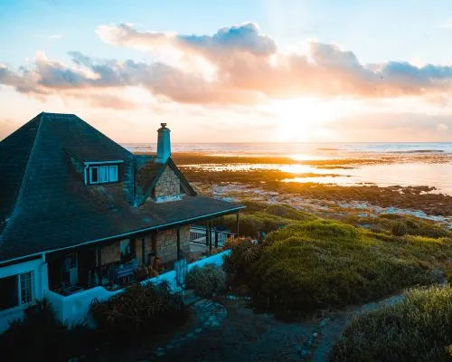 Coastal cottage with a blue roof overlooks a serene ocean sunset.