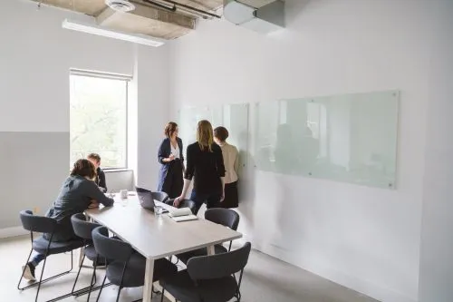 People collaborate in a meeting room, with some seated at a table and others standing near a whiteboard.