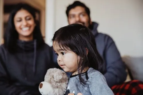 Young child holding a stuffed toy with two adults smiling in the background.