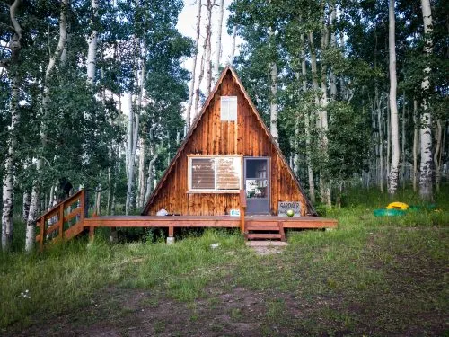 A-frame cabin surrounded by dense trees with a wooden porch and a small sign.