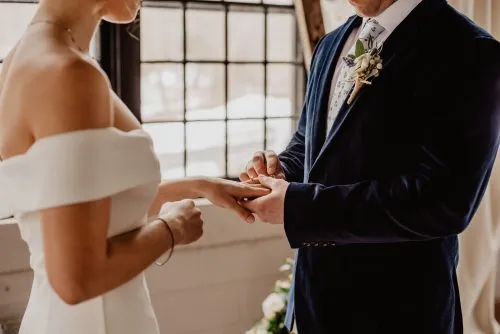 A couple exchanges rings during a wedding ceremony near a window.