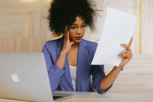 Woman in a blazer reviews a document at a desk with a laptop.