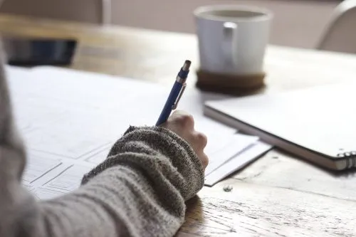 Person writing on paper at a wooden table with a coffee mug nearby.